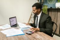 Seorang pengusaha muda tampak fokus menganalisis laporan keuangan di meja kerjanya, menunjukkan pentingnya perencanaan matang dalam mengelola modal usaha. Meja dipenuhi peralatan kantor dan laptop, mencerminkan aktivitas bisnis modern yang efisien.