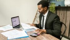 Seorang pengusaha muda tampak fokus menganalisis laporan keuangan di meja kerjanya, menunjukkan pentingnya perencanaan matang dalam mengelola modal usaha. Meja dipenuhi peralatan kantor dan laptop, mencerminkan aktivitas bisnis modern yang efisien.