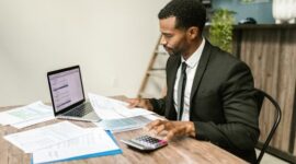 Seorang pengusaha muda tampak fokus menganalisis laporan keuangan di meja kerjanya, menunjukkan pentingnya perencanaan matang dalam mengelola modal usaha. Meja dipenuhi peralatan kantor dan laptop, mencerminkan aktivitas bisnis modern yang efisien.