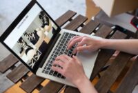 Seorang pemilik UMKM sedang mengoperasikan laptopnya di sebuah kedai kopi yang nyaman, menggambarkan dedikasi dalam mengelola pemasaran online produk lokalnya. Gambar ini merefleksikan bagaimana teknologi menjadi alat vital bagi pelaku usaha kecil untuk mengembangkan jangkauan pasar mereka.